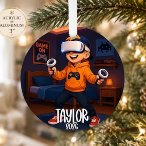 May include: A round Christmas ornament featuring a cartoon character wearing a VR headset and an orange hoodie, holding game controllers. The background shows a bedroom with a bed and a sign that says GAME ON. The name TAYLOR and 2026 are printed below.