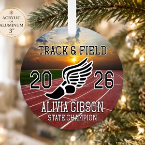 May include: A round 3-inch acrylic or aluminum ornament featuring a track and field graphic with a sunset over a running track. The text reads TRACK & FIELD, 2026, ALIVIA GIBSON, and STATE CHAMPION, with a black winged shoe icon in the center.