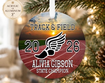 Puede incluir: Un adorno redondo de 7,62 cm de acrílico o aluminio con un diseño de atletismo sobre una pista al atardecer. El texto dice TRACK & FIELD, 2026, ALIVIA GIBSON y STATE CHAMPION, con un icono de zapato alado negro en el centro.