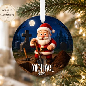 May include: A round 3-inch acrylic or aluminum ornament featuring a cartoon Santa Claus digging a grave in a cemetery at night under a full moon. The ornament includes the name Michael and the year 2026 printed in white text at the bottom.