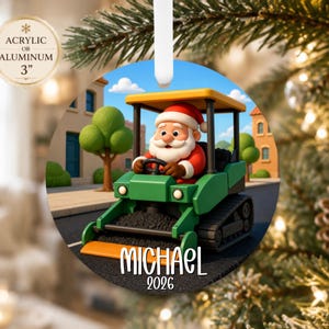 May include: A round 3-inch acrylic or aluminum ornament featuring a cartoon Santa Claus driving a green and yellow construction vehicle on a street. The name MICHAEL and the year 2026 are printed in white text at the bottom of the circular ornament.