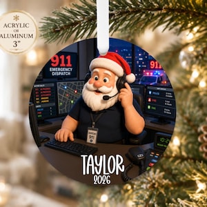 May include: A round 3-inch acrylic or aluminum ornament featuring a cartoon Santa Claus wearing a headset at a 911 emergency dispatch station with computer monitors, a keyboard, and a name tag reading Taylor 2026, hanging by a white ribbon.