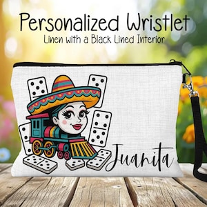 May include: White linen wristlet with a black zipper and wrist strap. The wristlet features a colorful illustration of a woman in a sombrero driving a train, surrounded by dominoes. The name "Juanita" is written in script.