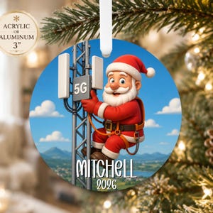 May include: A round Christmas ornament featuring a cartoon Santa Claus climbing a 5G cell tower. The ornament displays the name Mitchell and the year 2026 in white text. A circular label indicates the material is acrylic or aluminum and 3 inches.
