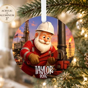 May include: A round acrylic or aluminum ornament featuring a cartoon Santa Claus in a white hard hat and red work uniform using a pipe wrench on an oil rig. The background shows a drilling rig at sunset with the text Taylor 2026 printed below.
