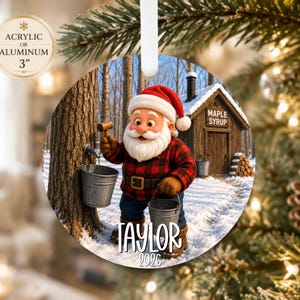 May include: A round 3-inch acrylic or aluminum ornament featuring a cartoon Santa Claus in a red and black plaid shirt, holding a mallet and metal bucket near a maple tree. A wooden cabin labeled MAPLE SYRUP is in the background. Text reads TAYLOR 2026.