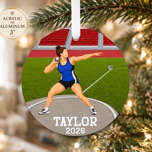 May include: A circular ornament featuring a shot put athlete in a blue and black uniform on a track and field throwing circle. The text TAYLOR 2026 is printed at the bottom. A small circular label indicates the material is acrylic or aluminum and 3 inches.