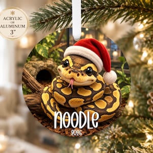 May include: A round 3-inch acrylic or aluminum ornament featuring a brown and yellow patterned snake wearing a red and white Santa hat. The snake has large black eyes and a pink tongue. The text NOODLE and 2026 is printed in white at the bottom.