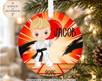 May include: A round acrylic or aluminum ornament measuring 3 inches in diameter. It features a cartoon illustration of a blonde child in a white karate uniform with a black belt. The background has red sunburst rays, black brush strokes, and the name JACOB with the year 2026 printed on it.