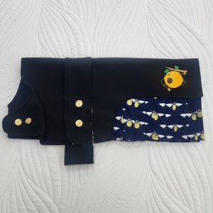 May include: A black dog coat with a beehive and bees design. The coat has gold buttons and a blue and white patterned lining.