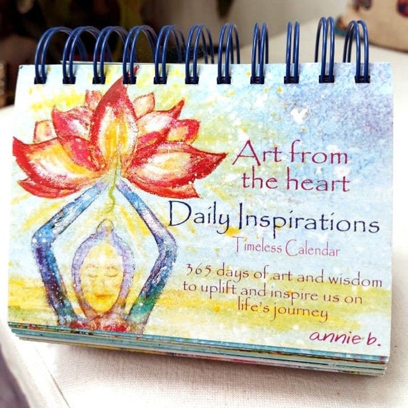May include: A colorful calendar with a watercolor painting of a person holding a lotus flower. The text on the cover reads "Art from the heart Daily Inspirations Timeless Calendar 365 days of art and wisdom to uplift and inspire us on life's journey annie b."