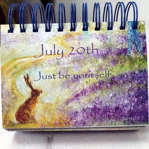May include: A watercolor painting of a hare sitting in a field of purple flowers. The painting has a light yellow background with a white swirl. The text "July 20th" and "Just be yourself" is written in purple on the painting.