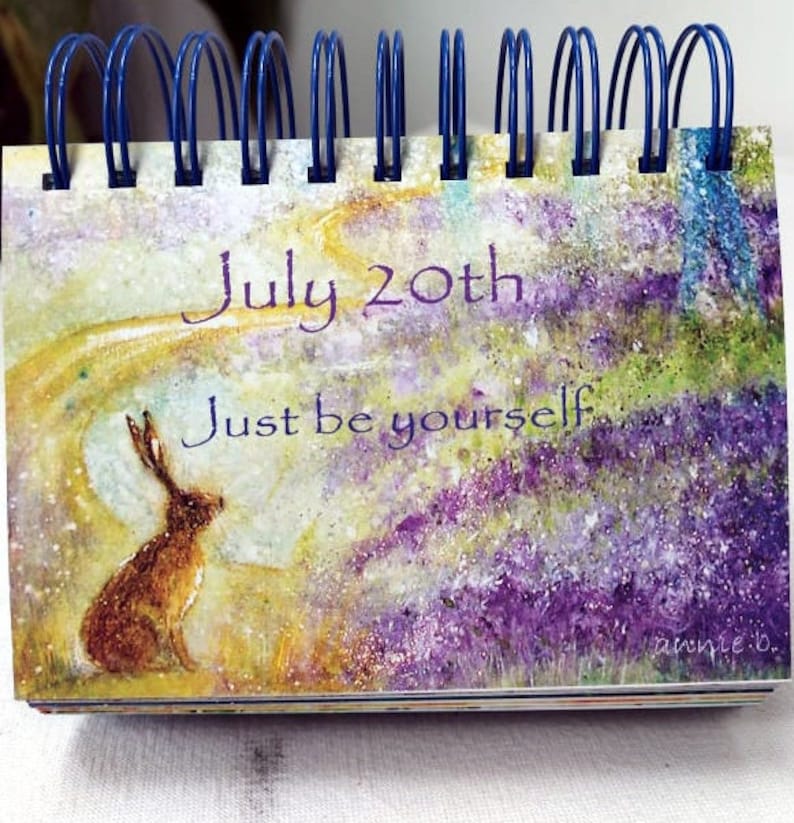 May include: A watercolor painting of a hare sitting in a field of purple flowers. The painting has a light yellow background with a white swirl. The text "July 20th" and "Just be yourself" is written in purple on the painting.