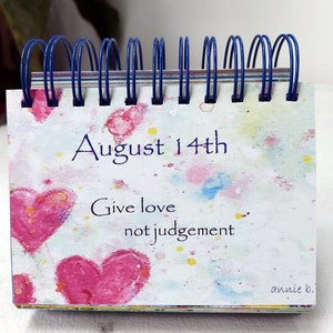 May include: A small desk calendar with a watercolor background featuring pink hearts and colorful splatters. The date is August 14th and the text reads "Give love not judgement". The bottom right corner is signed "annie b."