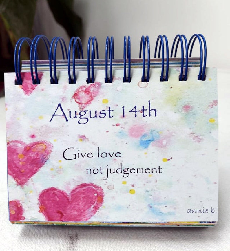 May include: A small desk calendar with a watercolor background featuring pink hearts and colorful splatters. The date is August 14th and the text reads "Give love not judgement". The bottom right corner is signed "annie b."