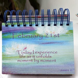 May include: A blue and white desktop calendar with the date February 21st. The calendar features a painted design of clouds and a quote that reads "Today I experience life as it unfolds moment by moment". The quote is signed "annie b."