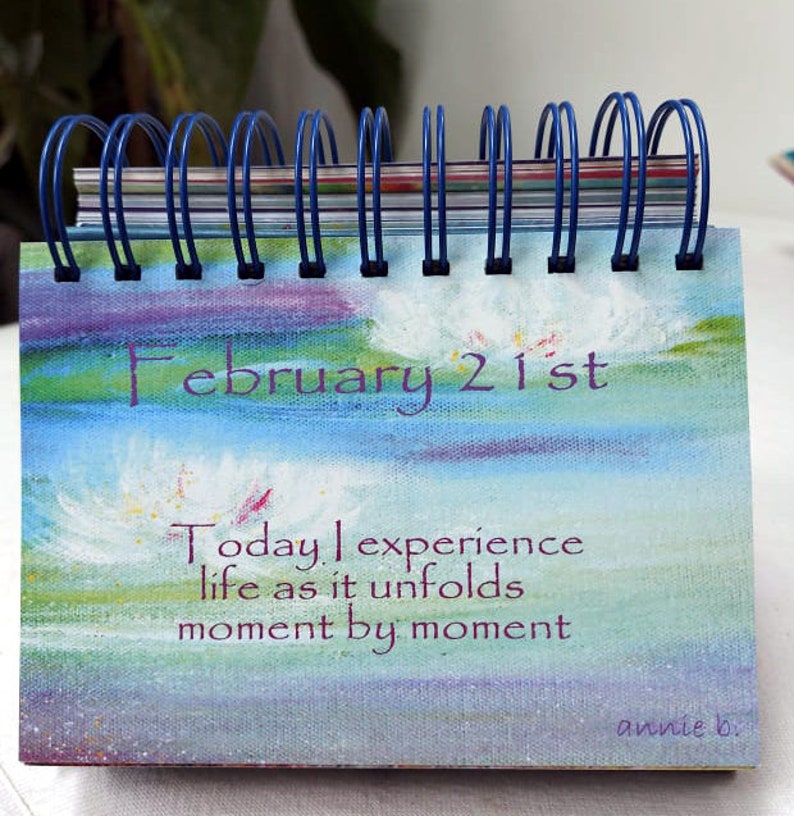 May include: A blue and white desktop calendar with the date February 21st. The calendar features a painted design of clouds and a quote that reads "Today I experience life as it unfolds moment by moment". The quote is signed "annie b."
