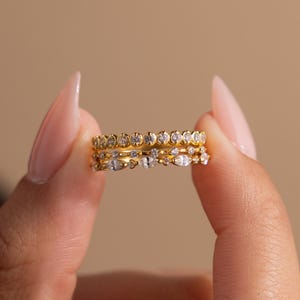 May include: Three gold rings with small, clear gemstones are held between fingers. The rings have different designs, with some featuring a row of stones and others with a mix of shapes. The background is a solid, neutral color.