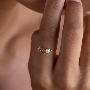 Puede incluir: Un anillo fino de oro con dos piedras pequeñas colocadas una al lado de la otra en una banda delicada. Una piedra es una perla blanca redonda y la otra es una piedra preciosa de color verde oliva alargada con corte marquesa.