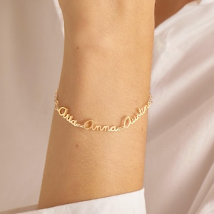 Puede incluir: Una pulsera de cadena en tono dorado con tres nombres conectados escritos en una fuente cursiva. Los nombres que aparecen en los delicados eslabones metálicos son Ava, Anna y Austin. La pulsera se lleva en la muñeca, mostrando las letras.