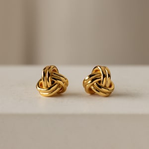 May include: A pair of gold knot-shaped earrings. The earrings are small and delicate, and they have a simple, elegant design.