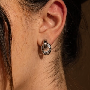 May include: Silver-toned stud earrings featuring a linked ring design. The earrings have a textured, metallic finish and are worn on the ear. The image shows a close-up of the ear and earring against a neutral background.