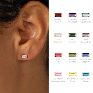 Może przedstawiać: Zbliżenie ucha z małym, prostokątnym kolczykiem z diamentem. Obraz przedstawia tabelę kolczyków z kamieniami szlachetnymi, z których każdy reprezentuje miesiąc urodzenia: Styczeń Granat, Luty Ametyst, Marzec Akwamaryn, Kwiecień Diament, Maj Szmaragd, Czerwiec Aleksandryt.