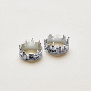 Puede incluir: Dos anillos de tono plateado con un diseño detallado de horizonte recortado. Las bandas de metal están grabadas con intrincadas siluetas arquitectónicas de edificios y rascacielos, creando un motivo de paisaje urbano continuo alrededor de cada anillo.