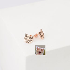 Puede incluir: Un par de pendientes de bot&oacute;n dorados con un dise&ntilde;o de cara de gato. Los pendientes se muestran sobre una superficie blanca.
