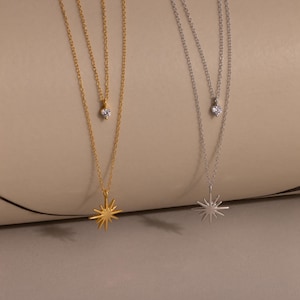 May include: Two layered necklaces, one gold and one silver, each with a small, clear gemstone pendant and a larger starburst pendant. The gold necklace has a sunburst pendant, while the silver necklace has a starburst pendant.