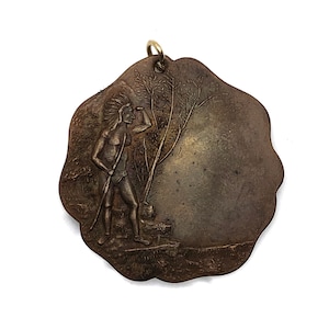 May include: A bronze-colored metal pendant featuring a raised image of a Native American man in a headdress, holding a spear and looking to the right. The pendant is shaped like a circle with a scalloped edge.