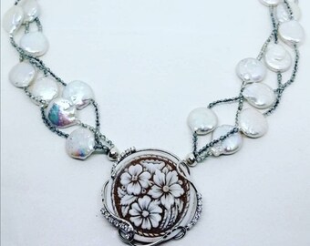 May include: A silver necklace with a white cameo pendant featuring a carved floral design. The necklace is made with multiple strands of small gray beads and white pearl-like discs.