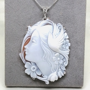 May include: A white cameo pendant necklace featuring a woman's profile with a dove and floral details. The pendant is set on a silver chain.