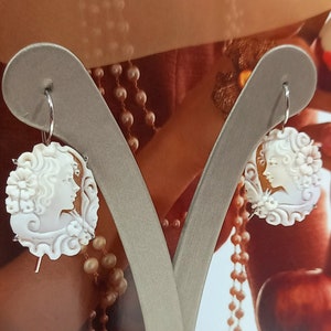 Puede incluir: Un par de pendientes de camafeo blancos con el perfil de una mujer en un diseño floral. Los pendientes están colgados de aros plateados y se exhiben en un soporte gris. El fondo borroso presenta perlas y una paleta de colores rojizos.