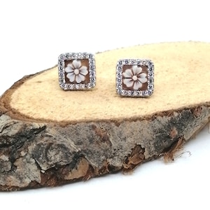 Puede incluir: Un par de pendientes cuadrados. Cada pendiente presenta un diseño de flor tallada en un tono marrón claro, rodeado por un borde de pequeñas piedras preciosas transparentes. Los pendientes están engastados en un metal de color claro.