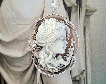 May include: A cameo pendant featuring a woman's profile with a floral headdress and a cherub. The cameo is set in a silver frame with a delicate diamond design.