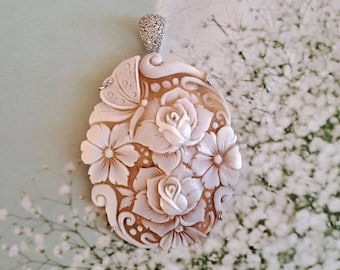 May include: A delicate, intricately carved shell cameo pendant featuring a floral design with roses and a butterfly. The cameo is set in a silver tone metal with a diamond-like accent at the top.