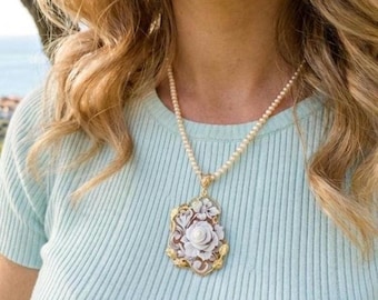 May include: A pearl necklace with a gold pendant featuring a white rose cameo. The pendant is framed by a gold filigree design. The necklace is worn over a light blue ribbed shirt.
