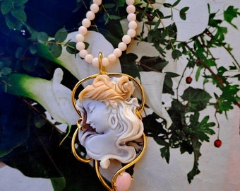 May include: A gold necklace with a pink shell cameo pendant featuring a woman's face. The pendant is surrounded by a gold frame and has a pink opal stone at the bottom.