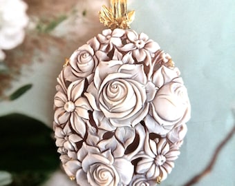 May include: A delicate white cameo pendant featuring a carved floral design with roses and other flowers. The pendant is set in a gold tone metal with a loop for attaching a chain.