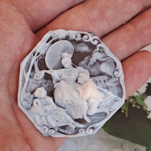 May include: A cameo brooch featuring a detailed carving of three figures, two women and a man, under an umbrella. The cameo is set in a dark brown, octagonal frame with intricate scrollwork.