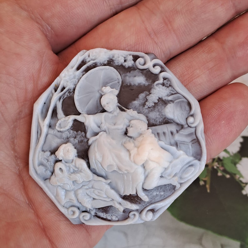 May include: A cameo brooch featuring a detailed carving of three figures, two women and a man, under an umbrella. The cameo is set in a dark brown, octagonal frame with intricate scrollwork.