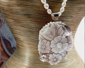 May include: A white pearl necklace with a carved shell pendant featuring a floral design. The pendant is made of white shell and has a silver tone finish.