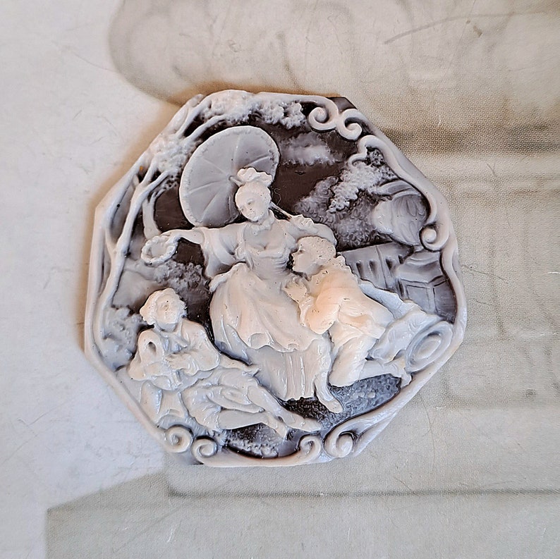 May include: A white cameo brooch with a detailed design of three figures in a garden setting. The figures are a woman holding an umbrella, a man, and a woman. The cameo is set in a white octagonal frame with a black background.