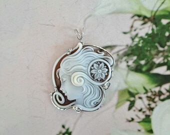 May include: A white cameo pendant featuring a carved portrait of a woman with a floral design. The pendant is set in a silver frame with a delicate floral design.