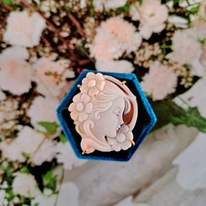 May include: A white cameo brooch with a detailed carving of a woman's face surrounded by flowers. The cameo is set in a gold tone frame and is displayed in a blue velvet box.