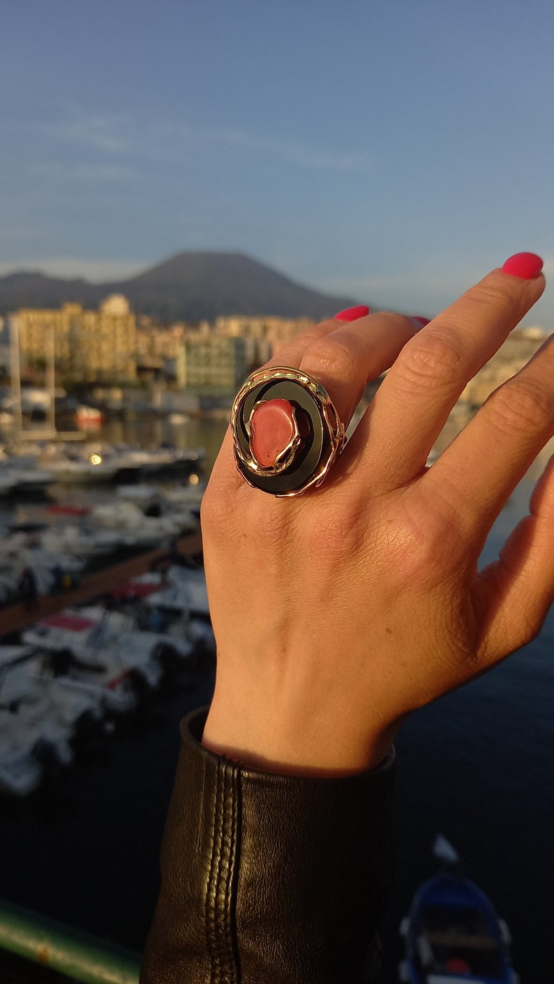 May include: A gold ring featuring a black and coral stone. The ring is worn on a person's finger.