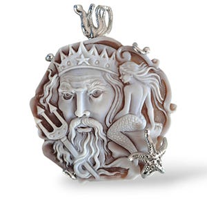 Puede incluir: Un colgante de camafeo de concha blanca y marrón con una talla detallada de Poseidón, el dios griego del mar, con un tridente y una sirena. El colgante está hecho de concha y plata.