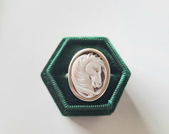 Puede incluir: Un anillo de camafeo blanco con un diseño de cabeza de caballo engastado en un engaste ovalado de tono dorado. El anillo se muestra en una caja de anillo de terciopelo verde.