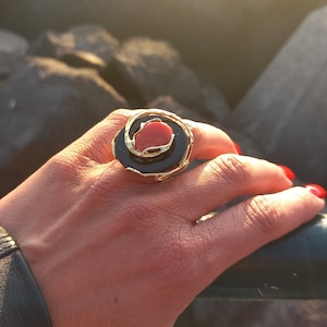 May include: A gold ring featuring a black and red stone. The ring is worn on a person's finger.